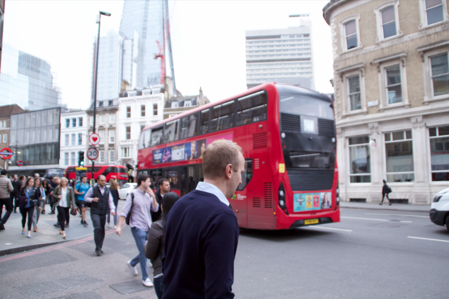 A Londoner motions to cross to the other sidewalk as a cherry red double decker rushes down the busy street.