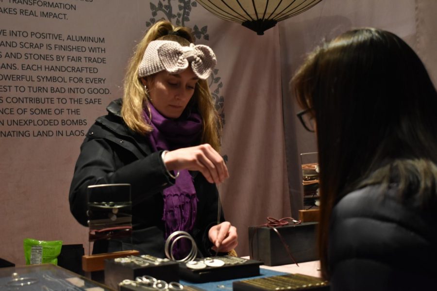 A kiosk employee dressed in a wool headband hands a silver necklace to a customer. This booth sold jewelry made from detonated bombs.
