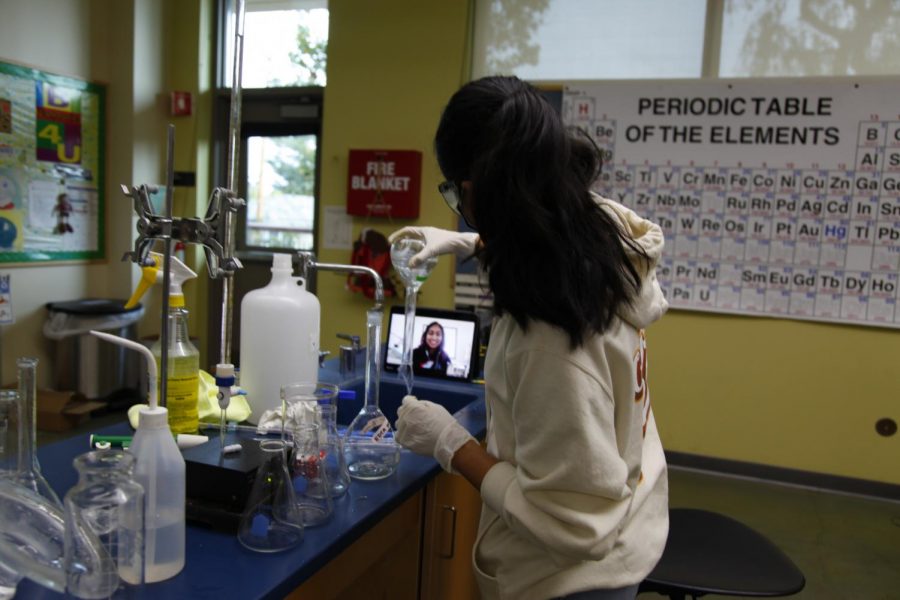 Nilisha Baid (12) pours out the contents of a volumetric flask into a sink during an experiment as her lab partner, Vaishnavi Murari (12), looks on via Zoom. Six out of the seven students in Honors Analytical Chemistry participated in the Nov. 21 in-person lab.