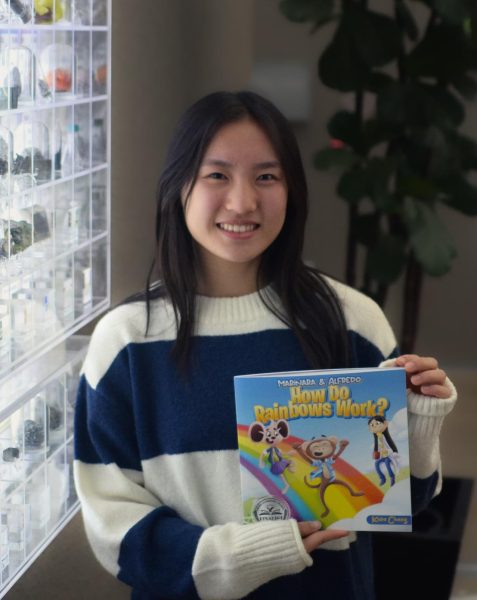 Keira Chang (10) holds her self-published children’s book titled “Marinara and Alfredo: How Do Rainbows Work?” next to the periodic table exhibit in Nichols Hall. “Growing up, I had a lot of questions about STEM and science in general, and I know other children are like that too,” Keira said.