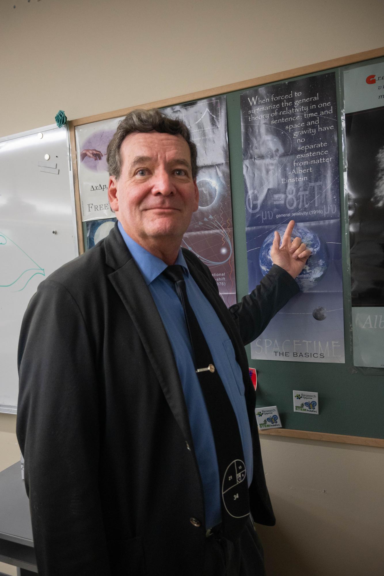 Dr. Eric Nelson points to a poster displaying Einstein's general relativity equation. Objects in his classroom range from NASA badges to a tesla coil.