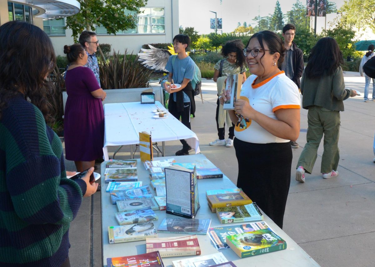 Assistant to Upper School Division Head Michelle Martinez holds up a book at Book Blog's table outside Nichols Hall. Martinez manages logistics for Cake Friday each month. 